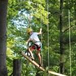 A camper crossing a low ropes balance element in the woods with spotters standing by on the Camp Akeela ropes challenge course