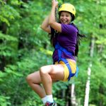 A camper in a helmet and harness riding the zip line down through the Vermont forest trees on the ropes course at Camp Akeela